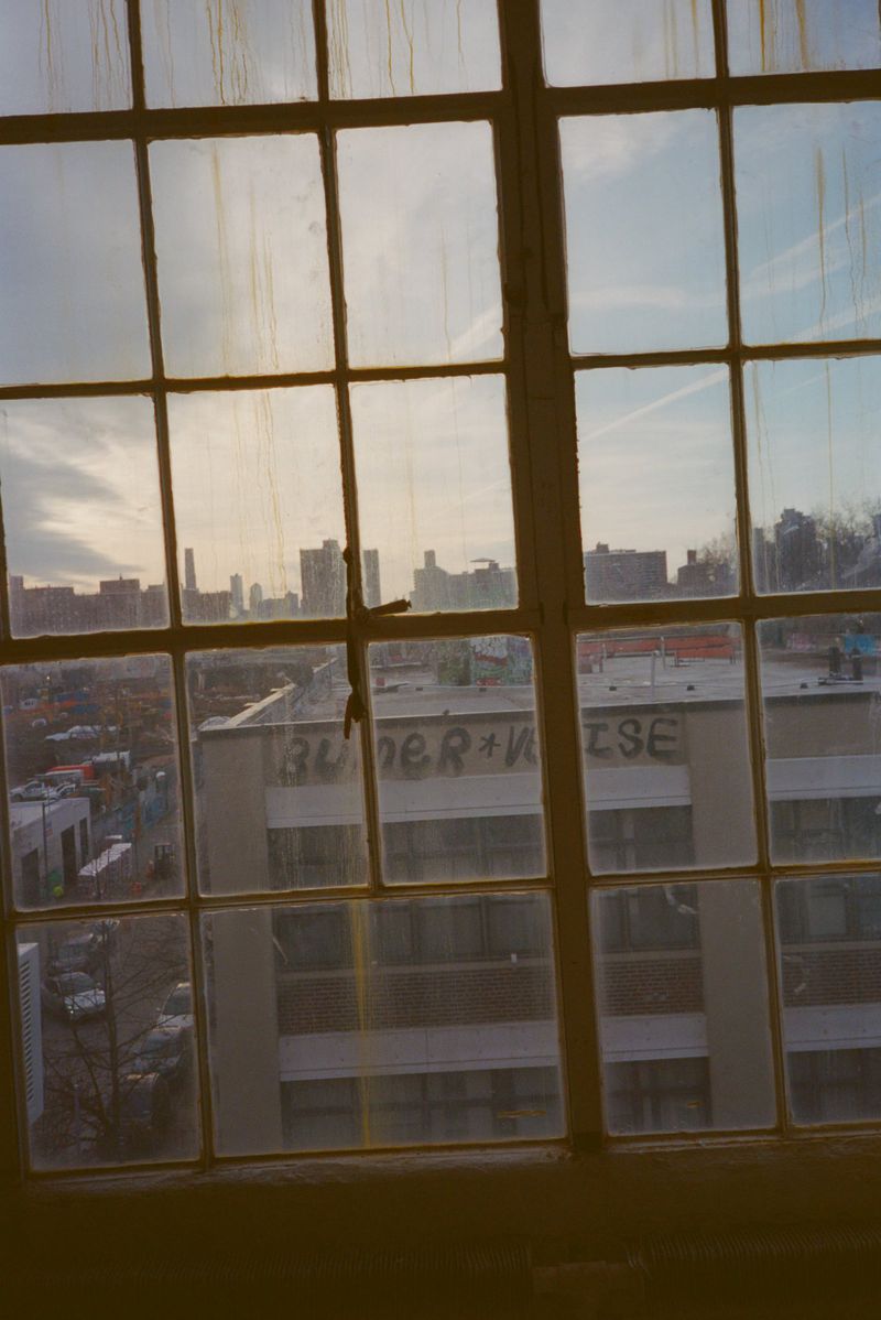 View of city buildings and rooftops seen through a large multi-pane industrial loft window in warm daylight.