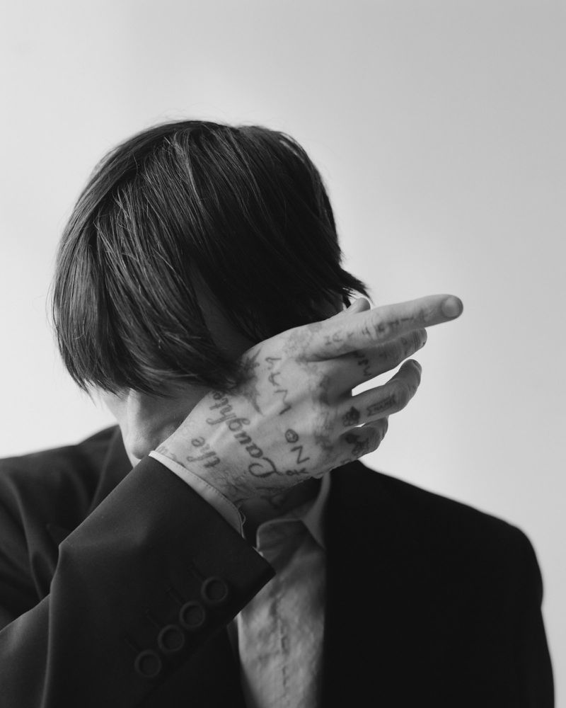 Model Cole Mohr covers his face with his tattooed hand while wearing a dark tailored blazer against a bright backdrop.