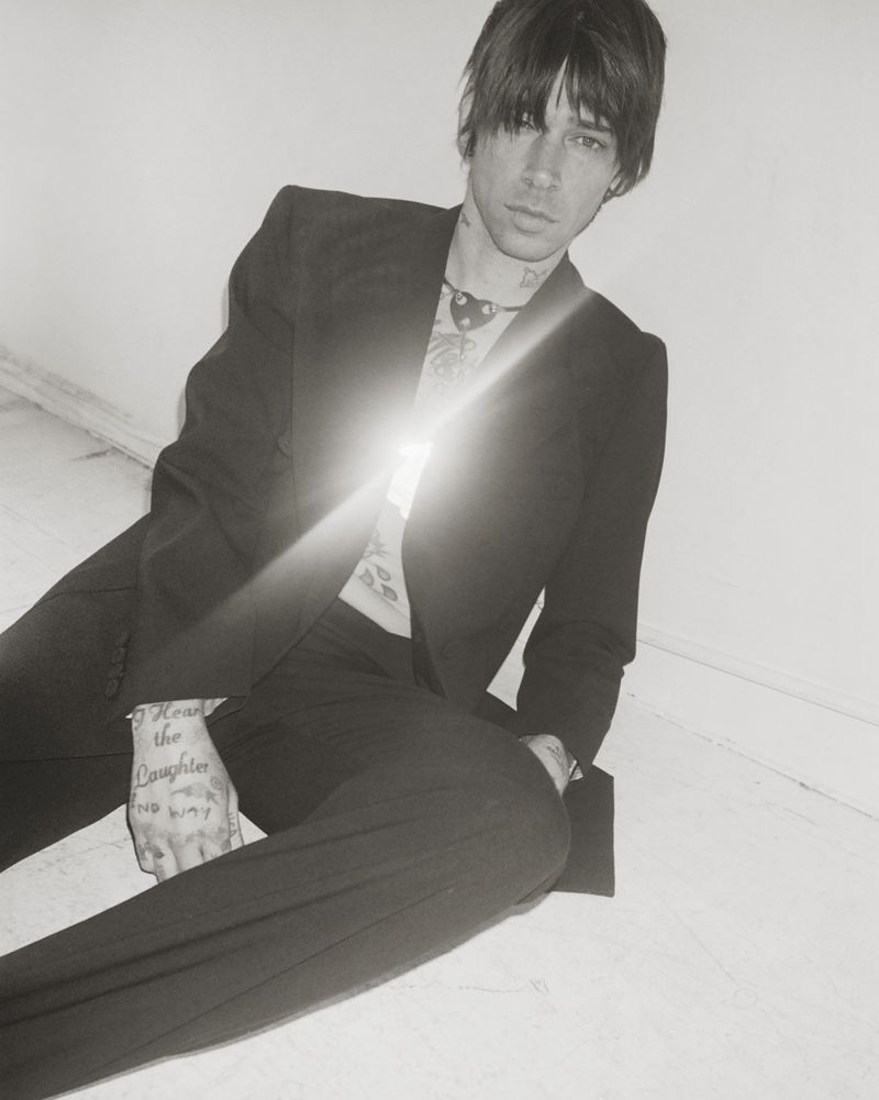Model Cole Mohr seated on the floor wearing an open black suit jacket and trousers in a black and white editorial portrait.