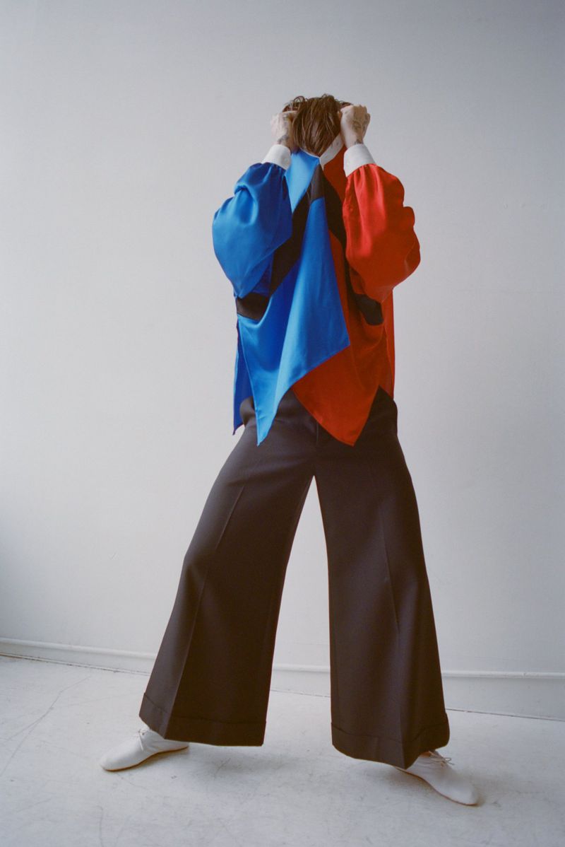 Cole Mohr posing in wide-leg dark trousers and a bright colorblocked blue and red tunic top against a white wall.