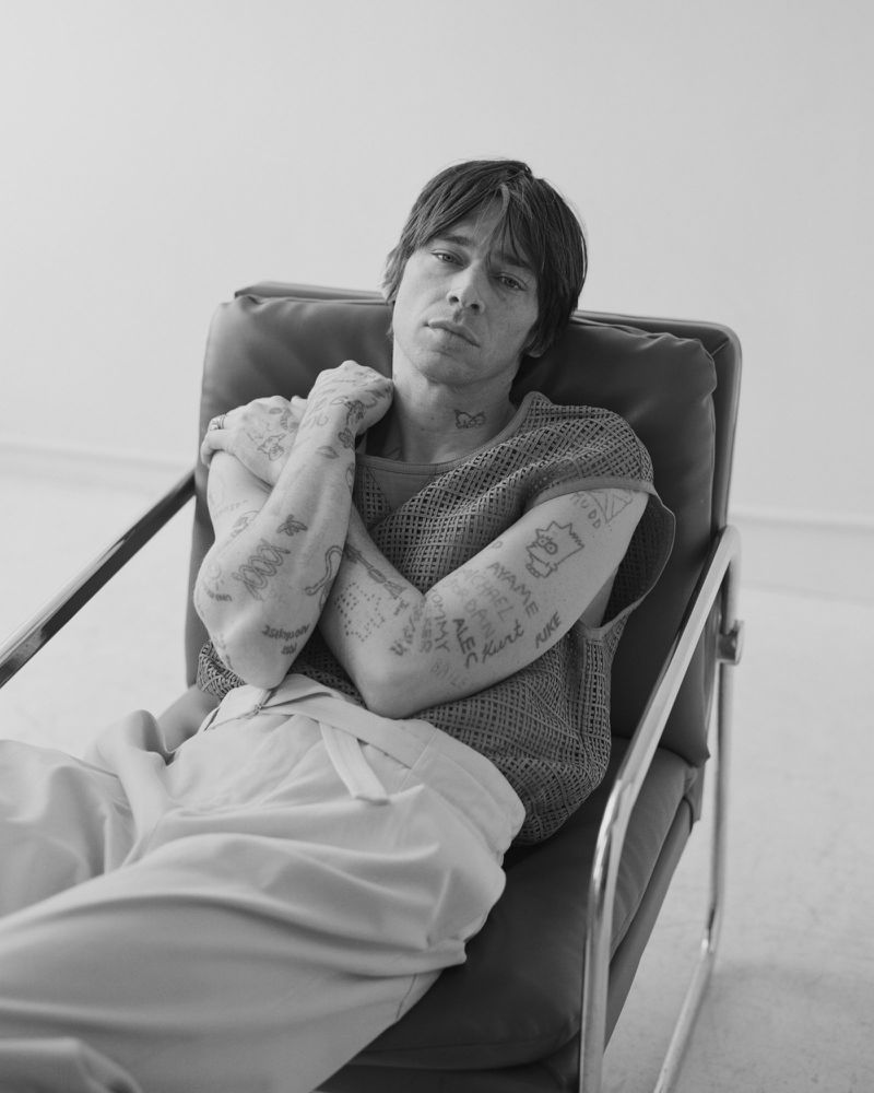 Cole Mohr with arm tattoos sitting in an armchair wearing a mesh top and trousers in a black and white studio portrait.