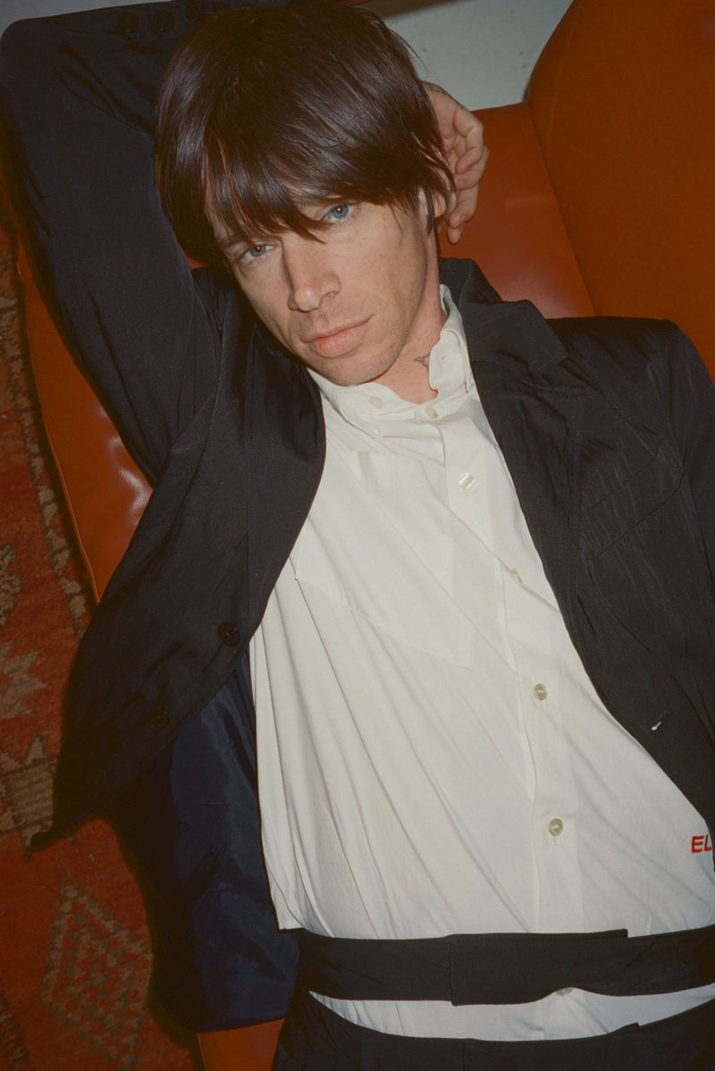 Model Cole Mohr reclining in a white button-up shirt and unbuttoned black jacket against an orange leather surface.