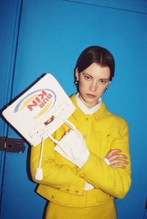Model posing in a bright yellow tweed jacket and white shirt holding a novelty graphic handbag against a blue backdrop.