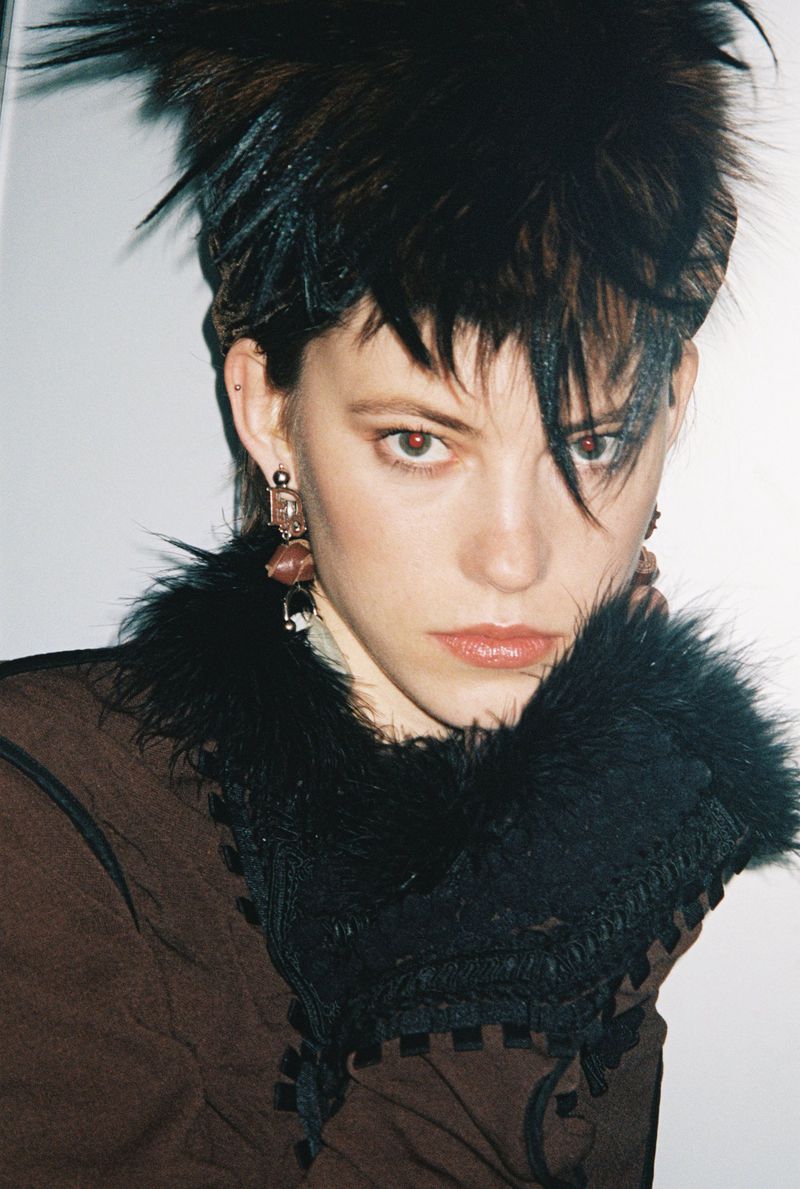 Close-up portrait of a model with spiky dark hair wearing a brown top with black feathered trim against a white background.