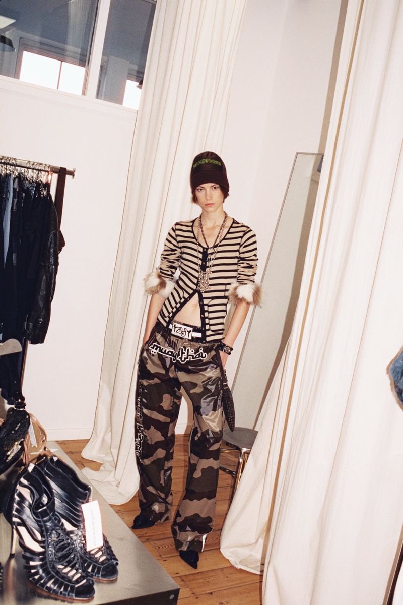 Model Dana Smith wearing a striped cropped cardigan, beanie, and camouflage pants posing in a backstage dressing area.