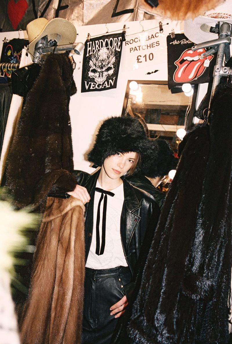 Model Dana Smith wearing a black fuzzy bucket hat and leather jacket surrounded by vintage fur coats on racks.