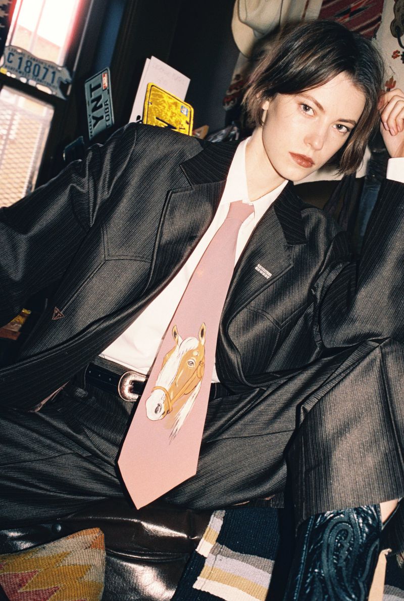 Model wearing a dark pinstripe suit and pink horse-printed tie in an interior setting for i-D Magazine