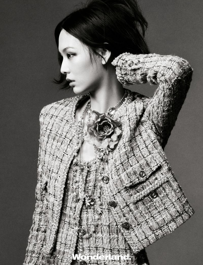 A woman poses wearing a tweed jacket, necklace, and matching tweed dress in a studio fashion shot against a gray background.