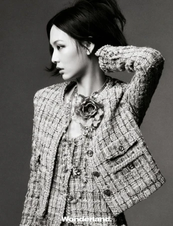 A woman poses wearing a tweed jacket, necklace, and matching tweed dress in a studio fashion shot against a gray background.