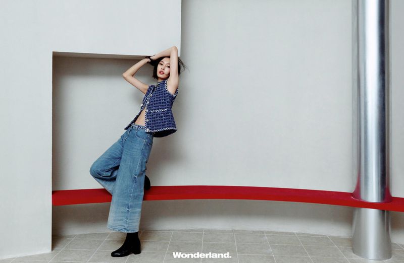 Adult woman in a sleeveless top and jeans posing against a white wall, with a metal pole and red bar adding a stylish backdrop.