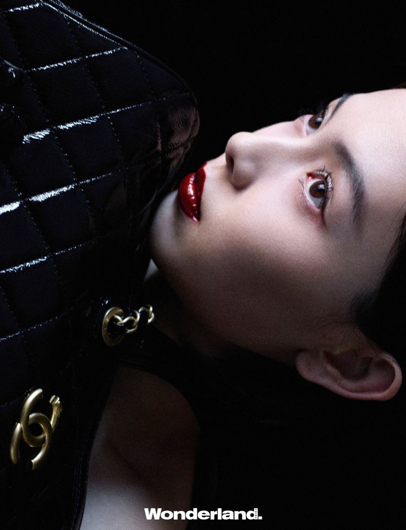 Close-up studio shot of a young adult woman with red lipstick looking up, with a glossy black quilted handbag and gold chain detail.
