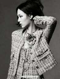 Black and white portrait of a woman wearing a textured tweed jacket and large floral necklace against a grey seamless background.