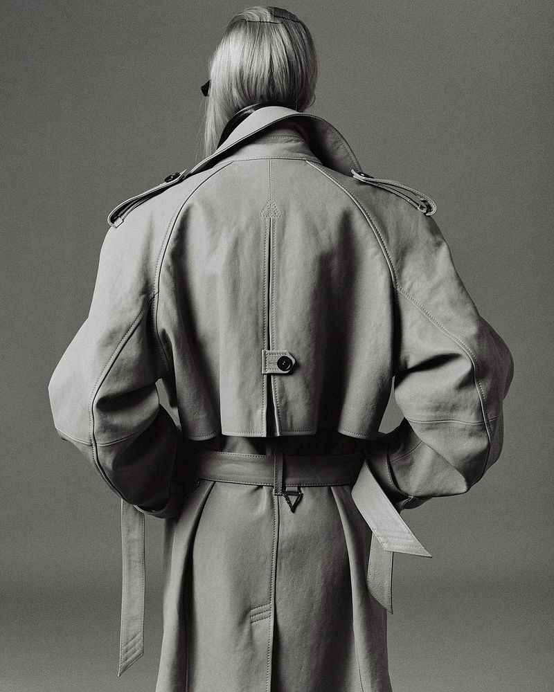 Model viewed from behind wearing an oversized belted trench coat with storm flap detailing against a neutral studio backdrop.