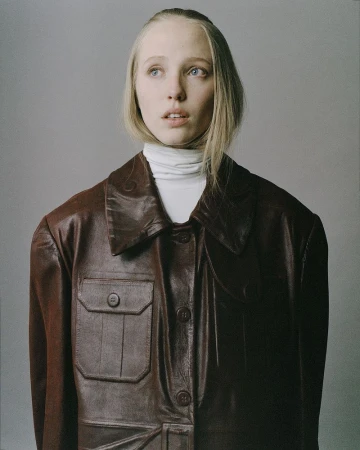 Model Maelle Swaelens wearing an oversized dark brown leather jacket and white turtleneck against a grey studio backdrop.