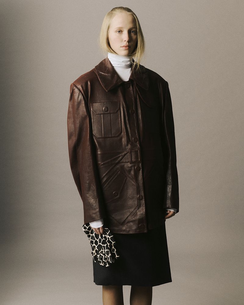 Model Maelle Swaelens wearing an oversized brown leather jacket, white turtleneck, and patterned glove against a grey studio backdrop.