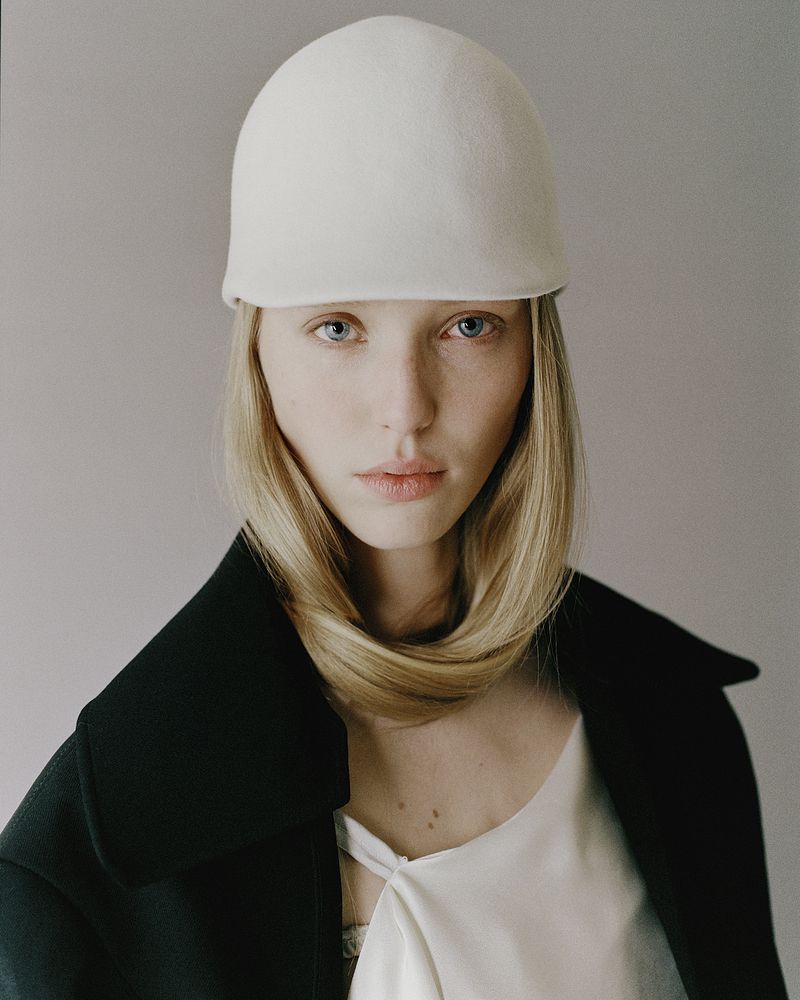 Model Maelle Swaelens wearing a sculptural white dome hat and a black coat against a neutral grey studio backdrop.