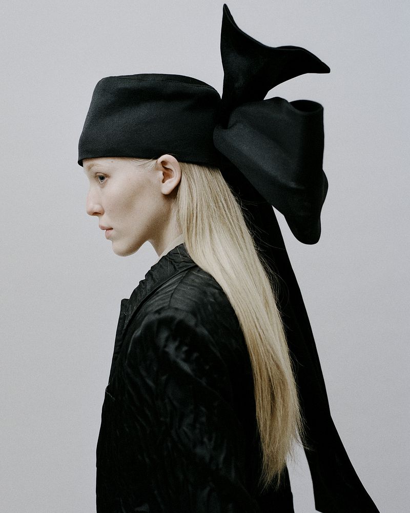 Model Maelle Swaelens in profile wearing a large black structured bow headpiece and black textured jacket against a pale backdrop.