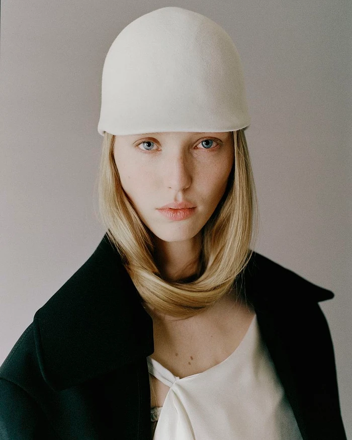 Model Maelle Swaelens wearing a sculptural white dome hat and a black coat against a neutral grey studio backdrop.