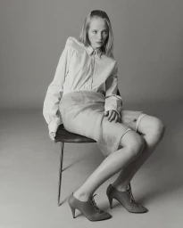 Model seated on a chair wearing a long-sleeve white button-up shirt, sheer skirt, and lace-up heels against a grey studio backdrop.