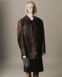 Model Maelle Swaelens wearing an oversized brown leather jacket, white turtleneck, and patterned glove against a grey studio backdrop.