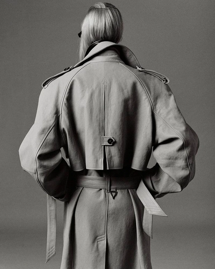 Model viewed from behind wearing an oversized belted trench coat with storm flap detailing against a neutral studio backdrop.