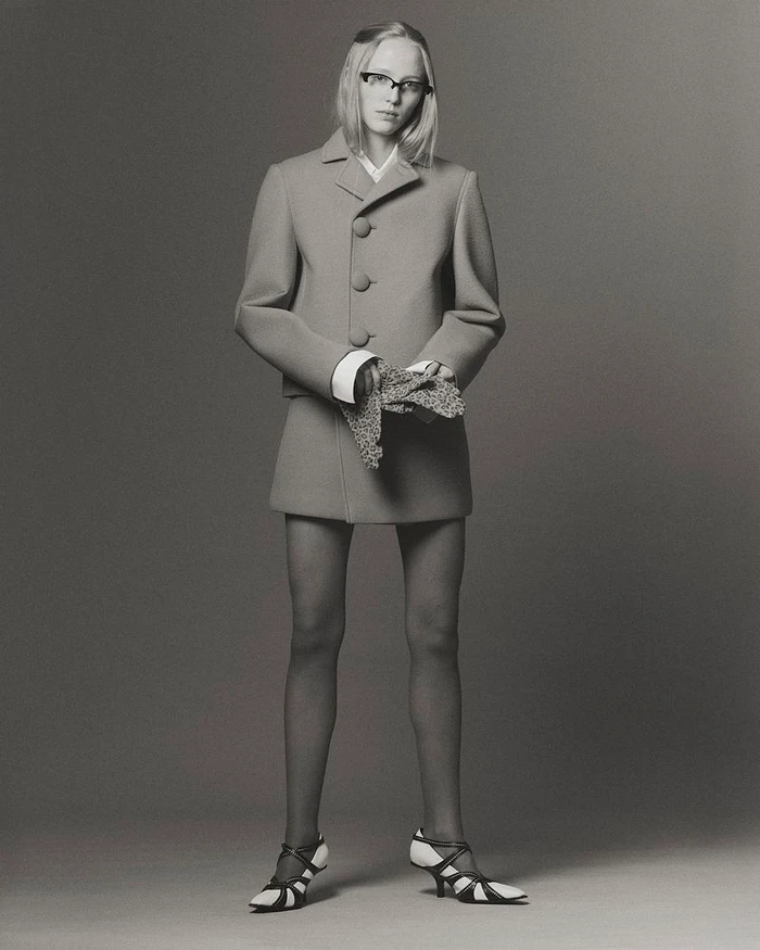 Model Maelle Swaelens posing in a structured button-up coat dress, dark tights, and strappy heels against a grey studio backdrop.