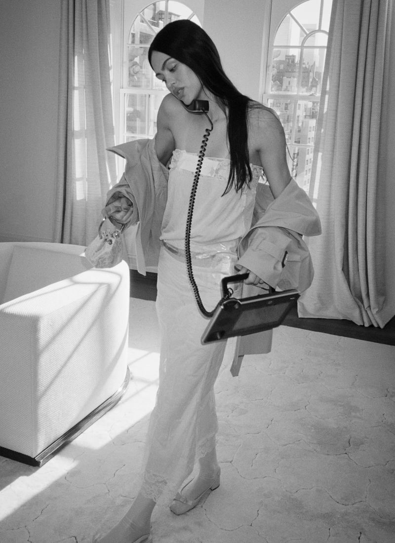 Monochrome shot of Amelia Gray wearing a strapless slip dress and trench coat while holding a corded telephone in a sunlit room.