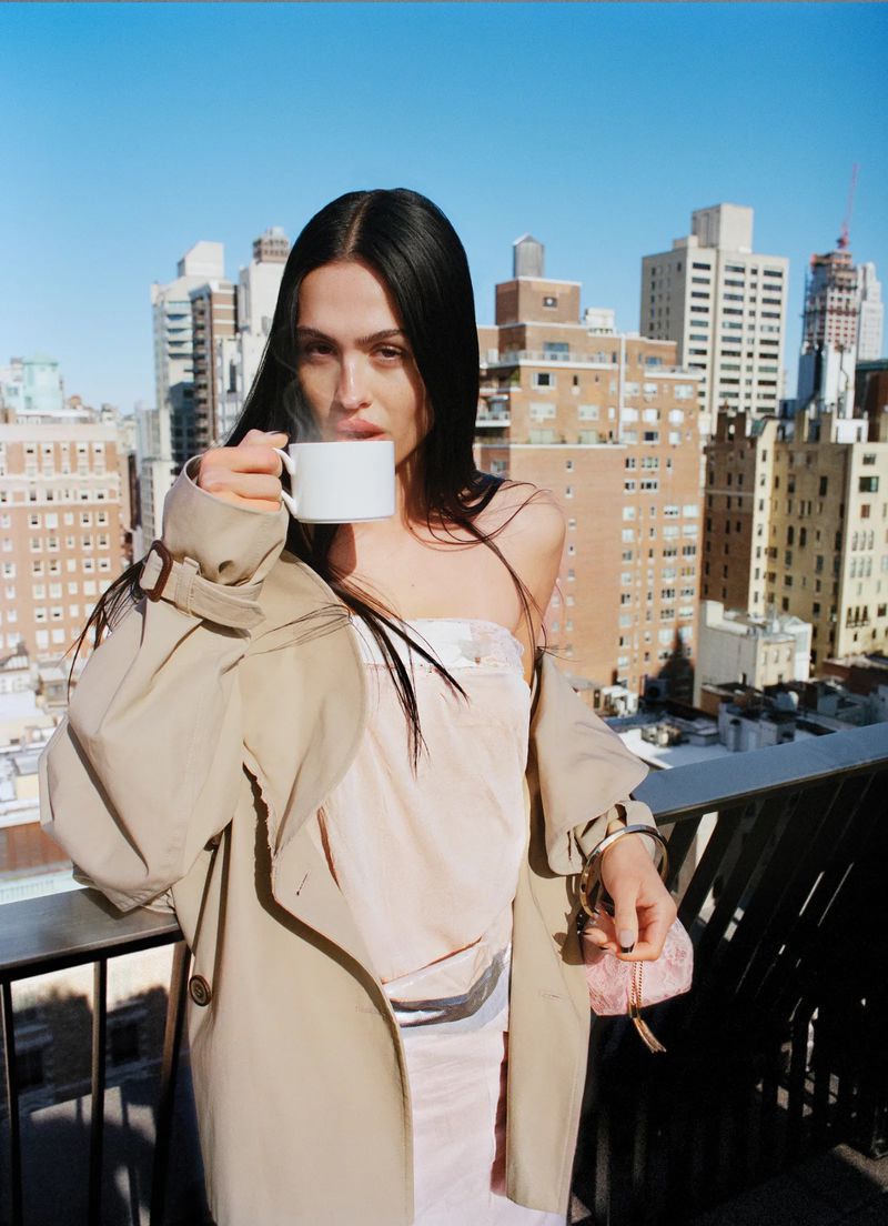 Model Amelia Gray drinking from a white cup on a city balcony wearing a beige trench coat over a pale dress.