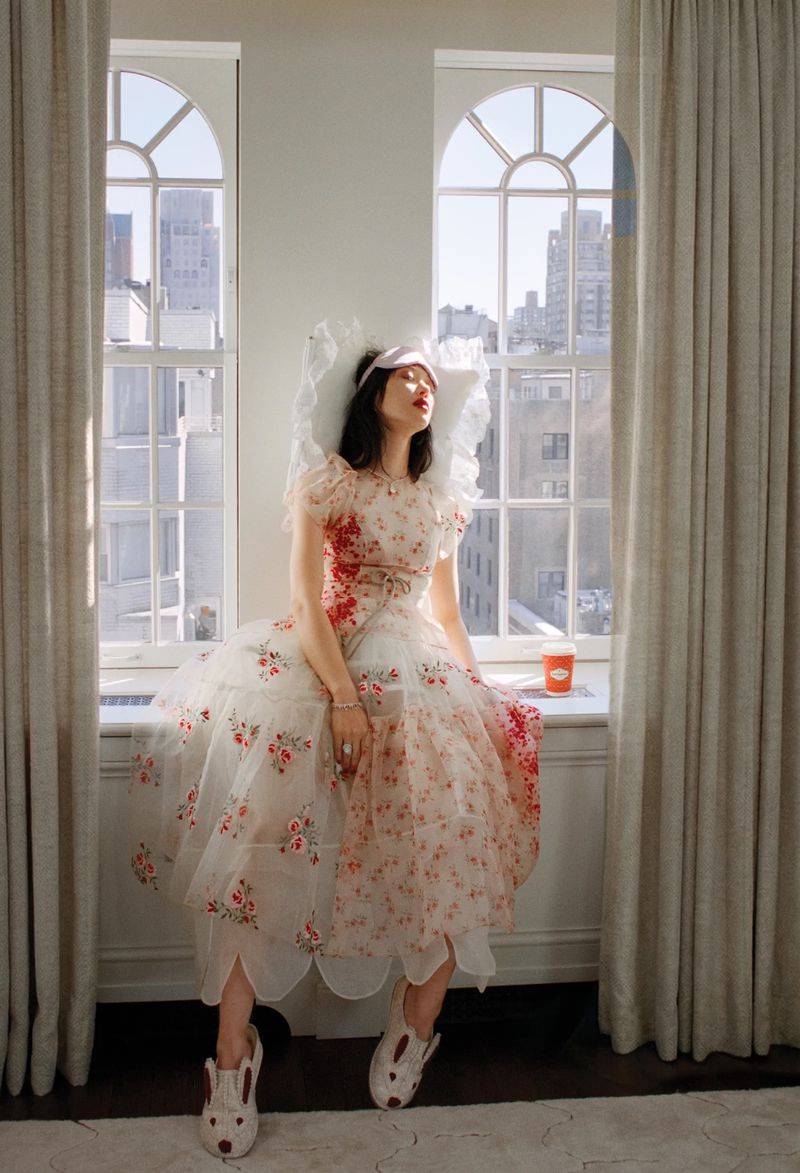 Sora Choi wearing a floral tulle dress and sleep mask while leaning on a white pillow on a windowsill between arched windows.
