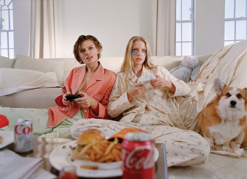 Two women in pajamas sit on a living room floor playing video games beside a corgi, with fast food and soda cans on the table.