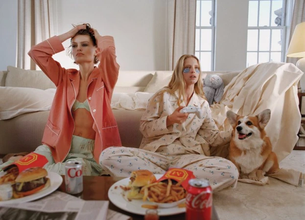 Two models in pajama sets sit on a living room floor beside a corgi dog with burgers, fries, and soda cans on a coffee table.