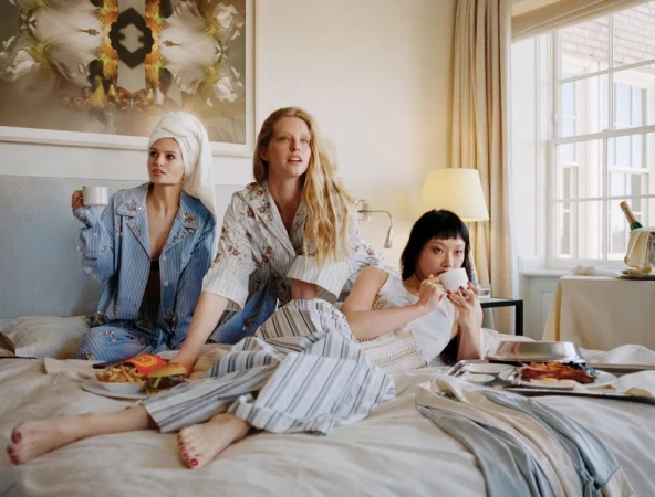 Three models lounging in striped pajamas on a hotel bed with room service trays and teacups.