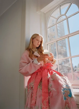 Model in a belted pink floral dress and jacket holding a cup while seated by a large arched window overlooking city buildings.