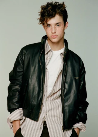 Young man with curly hair and freckles wearing a black leather jacket over a striped button-down shirt against a studio backdrop.