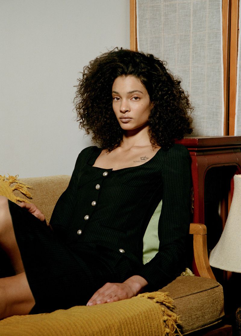 Woman with voluminous curly hair in a black square-neck buttoned dress sitting on a yellow upholstered armchair indoors.