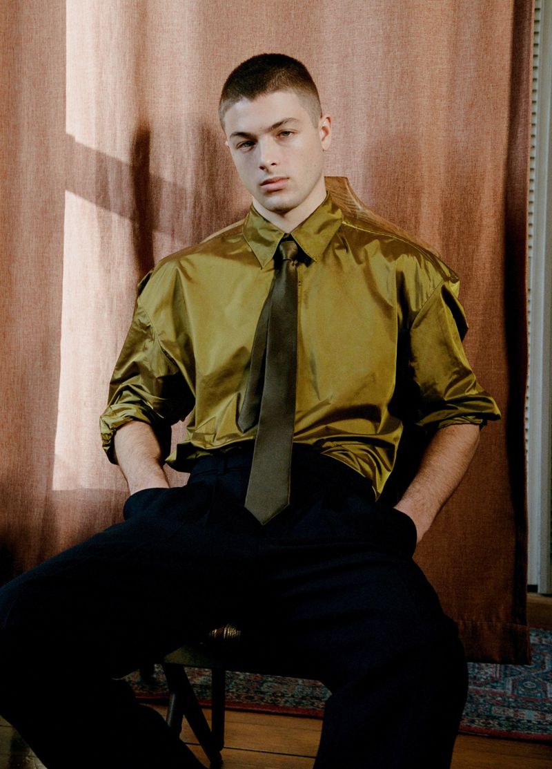 Male model seated in an olive green satin button-up shirt, olive tie, and dark trousers against draped brown curtains.