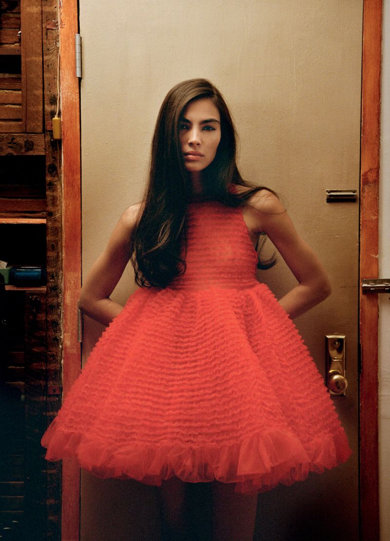 Model posing in a voluminous coral red ruffled babydoll dress against a neutral interior door frame.
