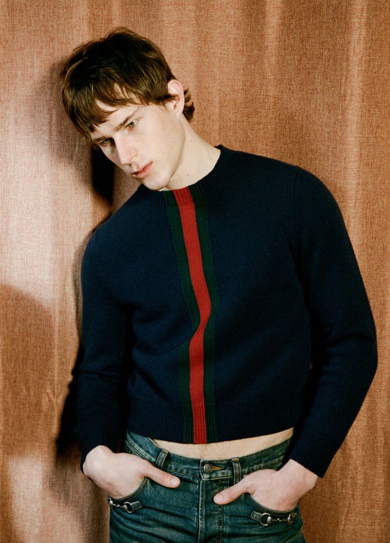 Young male model posing in a navy cropped sweater with green and red center stripe and blue jeans against a brown curtain.