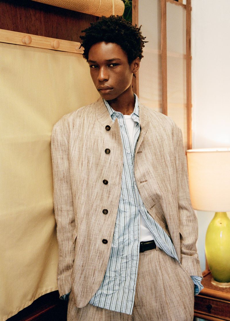 Young Black man in a textured beige button-up jacket and blue striped shirt standing beside a green table lamp.