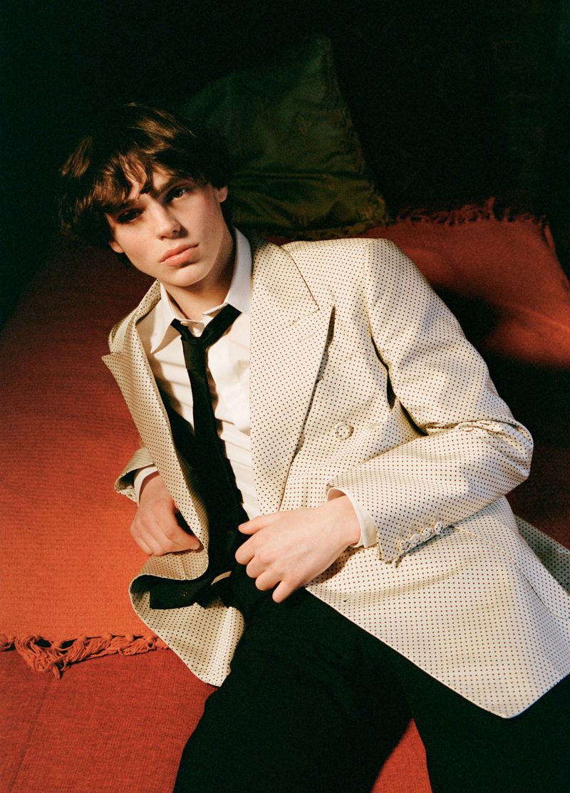 Young man in a cream patterned double-breasted blazer and black tie lying on a red bed for Hero USA