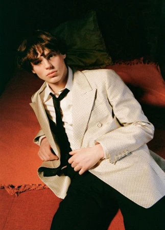 Young man in a cream patterned double-breasted blazer and black tie lying on a red bed for Hero USA