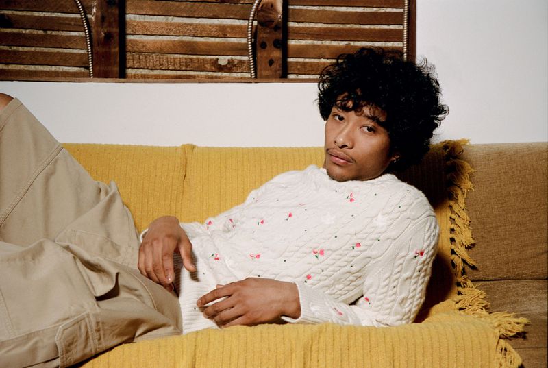 Man with curly dark hair reclining on a yellow sofa wearing an embroidered white cable knit sweater and beige cargo pants.