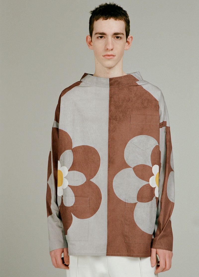 Young man wearing a brown and grey patterned floral long sleeve top against a solid neutral studio backdrop