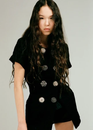 Model with long wavy dark hair wearing a black short-sleeve garment with ornate round buttons against a neutral studio backdrop.