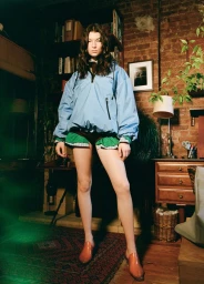 Model standing indoors wearing a light blue zip-up windbreaker, green trimmed shorts, and patent mules against a brick wall.