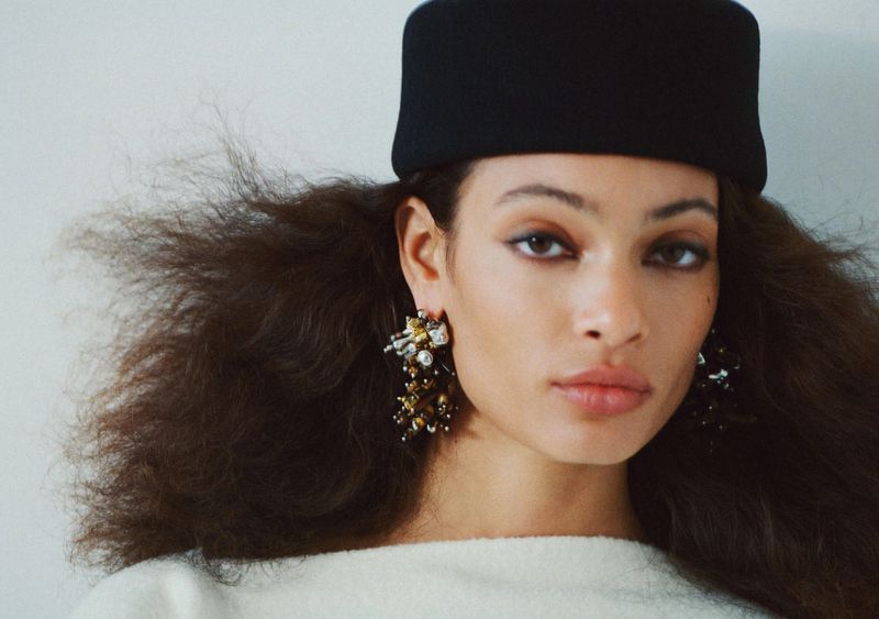 Close-up portrait of a woman with brown hair, wearing a black hat and statement earrings, against a plain white background.