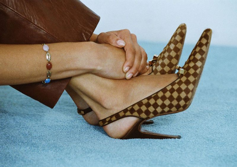 Close-up of a model wearing brown checkered slingback heels and a stone bracelet on light blue carpeting.