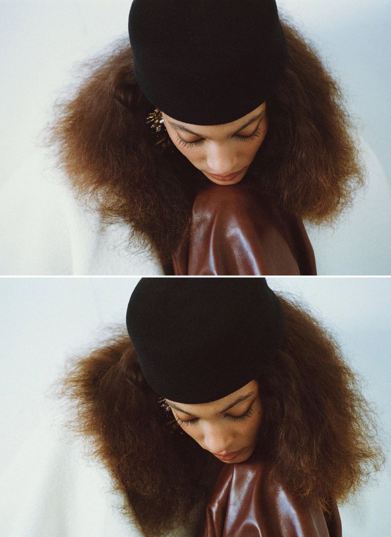 Model Aliza Jarmon looking down wearing a black cap, voluminous crimped hair, and brown glossy leather against a pale backdrop.