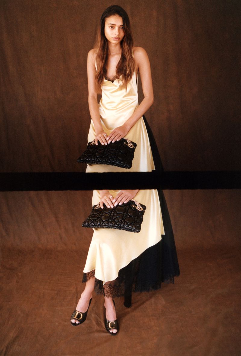 Model Jacoby Sorapuru posing in a cream satin slip dress with lace trim, holding a black quilted clutch against a warm brown backdrop.