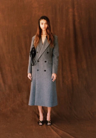 Model Jacoby Sorapuru stands in a long grey double-breasted coat and black heels against a textured brown studio backdrop.
